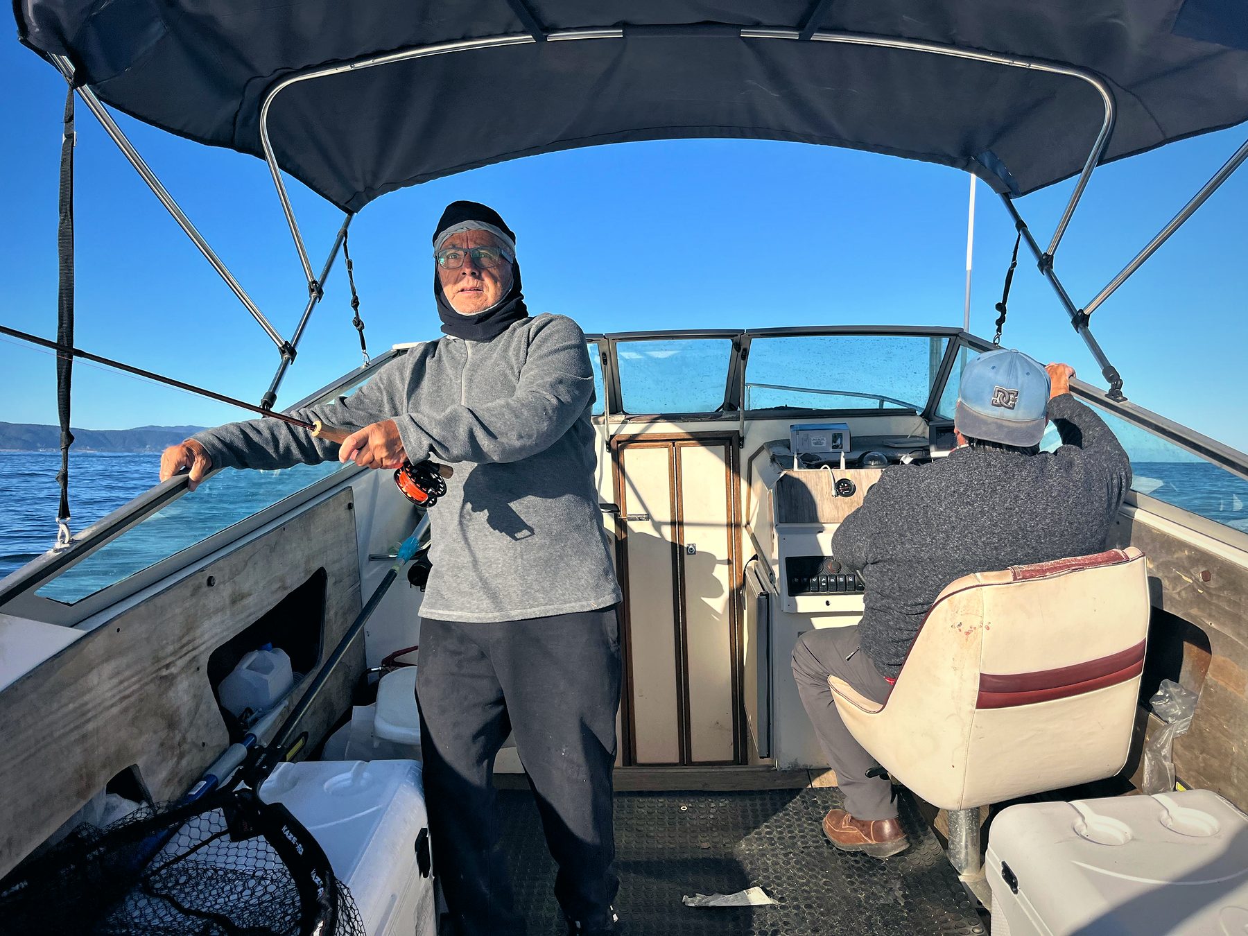 A warmly dressed angler with rod and reel on board under the canopy, blue sea behind and a crew member at the helm