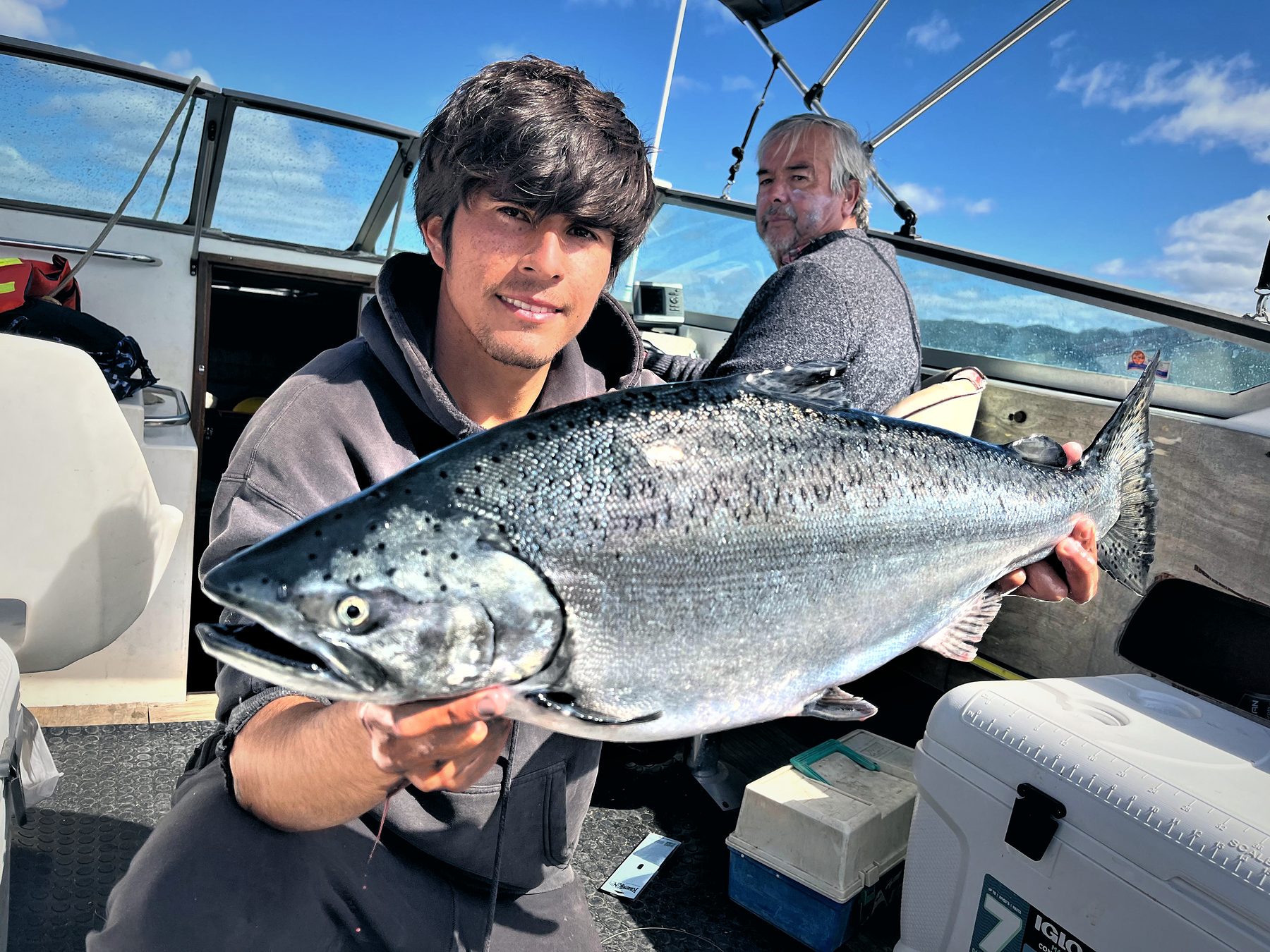 An angler smiles holding a silver chinook salmon on board, another crew member behind him and a blue sky