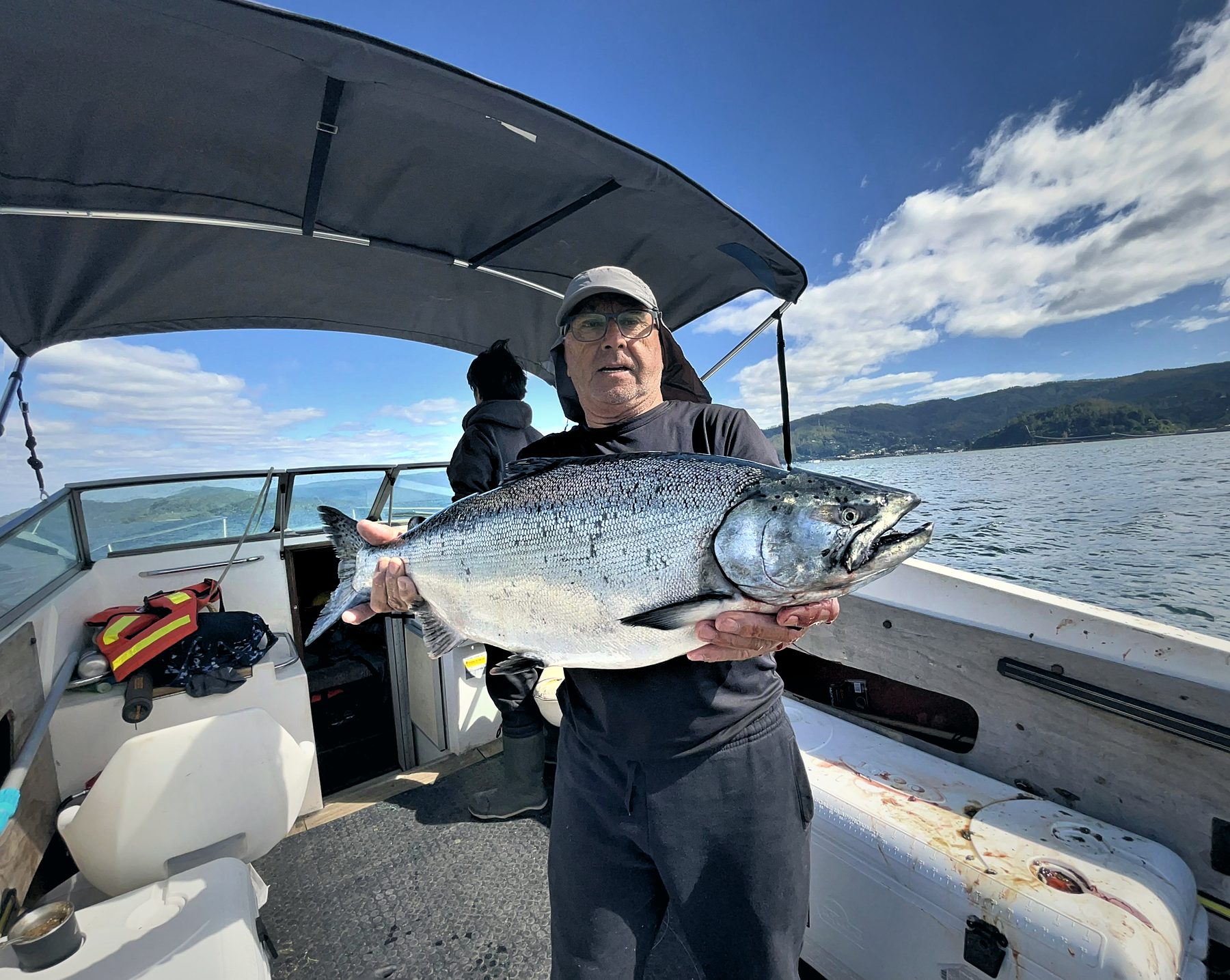 An angler standing at the stern holding a chinook salmon, with the sea and the coast behind